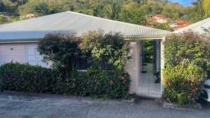 A single-story house with a corrugated metal roof, surrounded by lush greenery and a hillside in the background. - LILI Apartment with swimming pool, 400m from the beach. (Les Trois-Îlets)