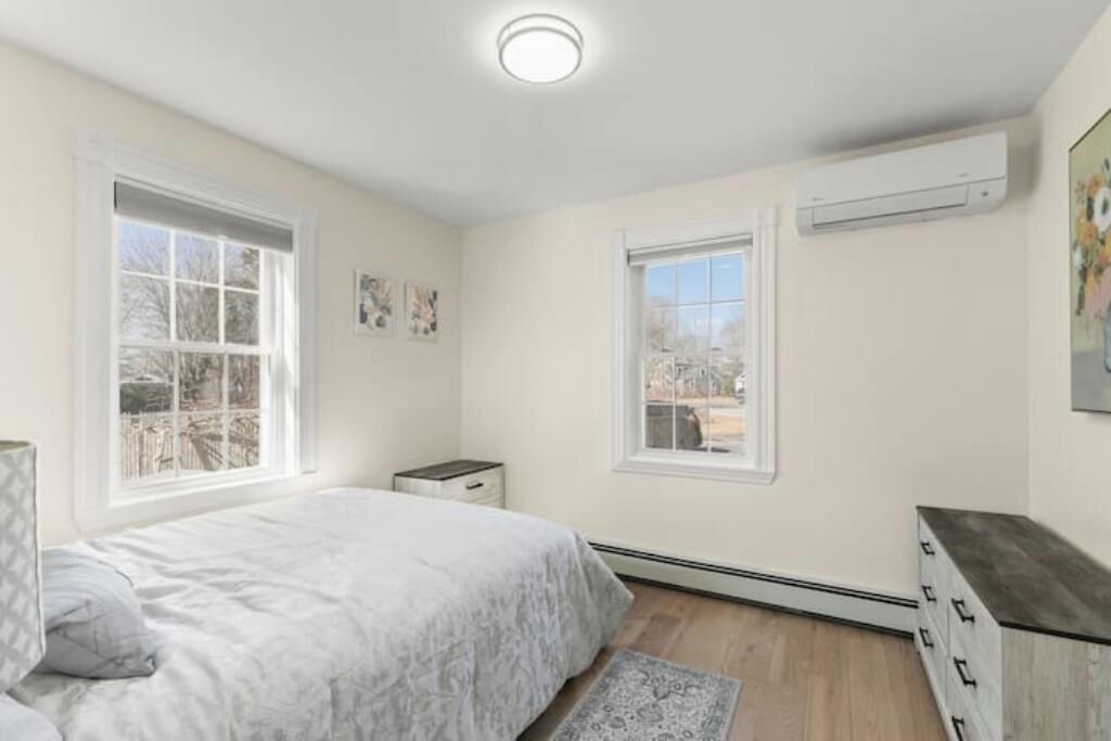Quarto com cama, duas janelas, ar condicionado, cômoda e mesa de cabeceira.