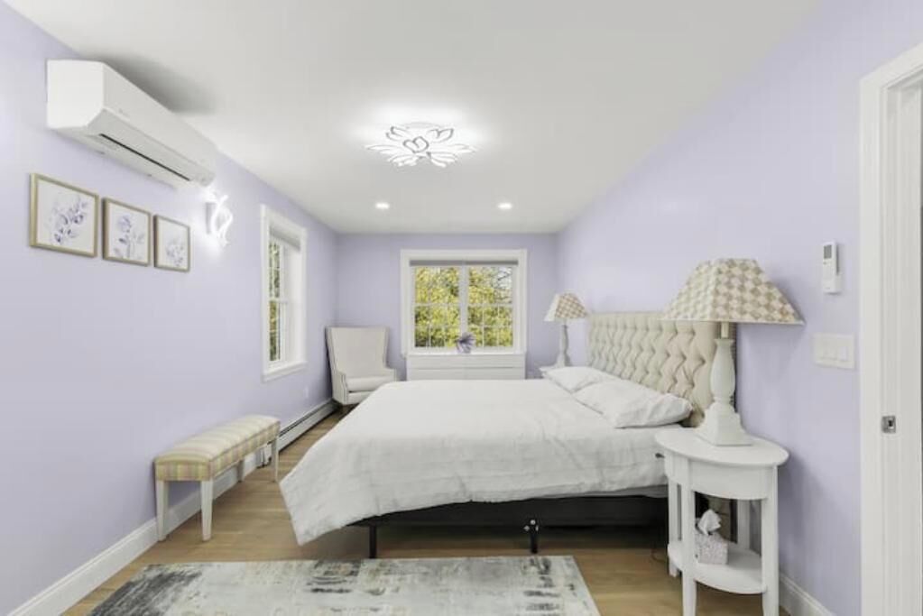 A bedroom with a bed, a bedside table, a chair, a window, and an air conditioning unit.