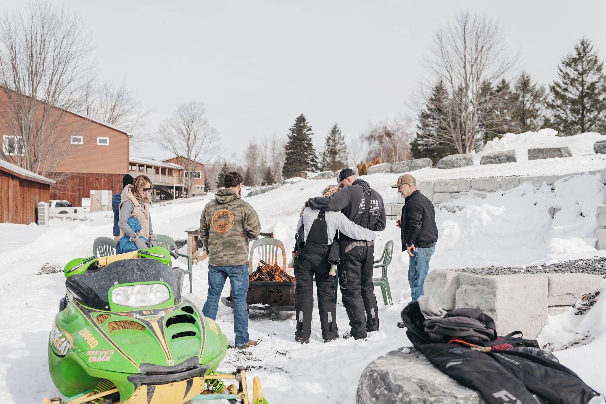 Un groupe de personnes s’est rassemblé autour d’un motoneige et d’un foyer dans un décor enneigé en plein air.