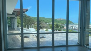 Un'ampia stanza con grandi vetrate scorrevoli che si aprono su una terrazza esterna con mobili bianchi e vista su un porto turistico e delle colline.