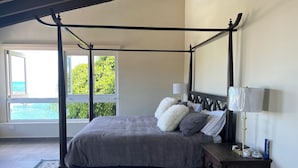A four-poster bed with a grey bedspread and pillows, a nightstand with lamps, a patterned rug, and a view of the sea through large windows. - Stunning ocean front villa with pool and power generator. 10min drive to beach (Fajardo)