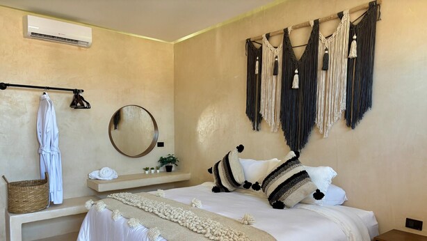 A bedroom with a bed, a bedside table, a hanging robe, a mirror, a wall-mounted air conditioner, and macramé wall hangings. - Ananda Hotel Boutique (Isla Holbox)