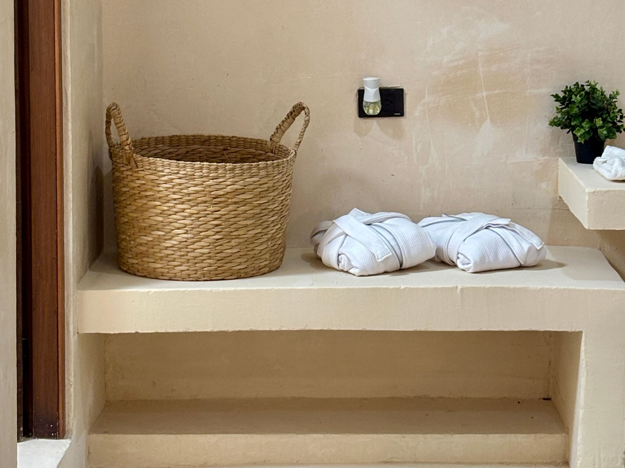 Een badkamer met een gevlochten mand, twee opgerolde handdoeken, een klein flesje en een potplant.