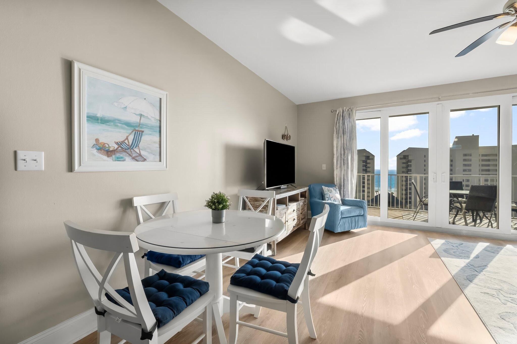 Un salon moderne avec une table ronde, des chaises blanches, un téléviseur et un balcon offrant une vue sur les immeubles.