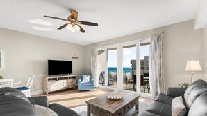 A living room with a television, a ceiling fan, a sofa, a coffee table, and a view of the ocean through large sliding doors.