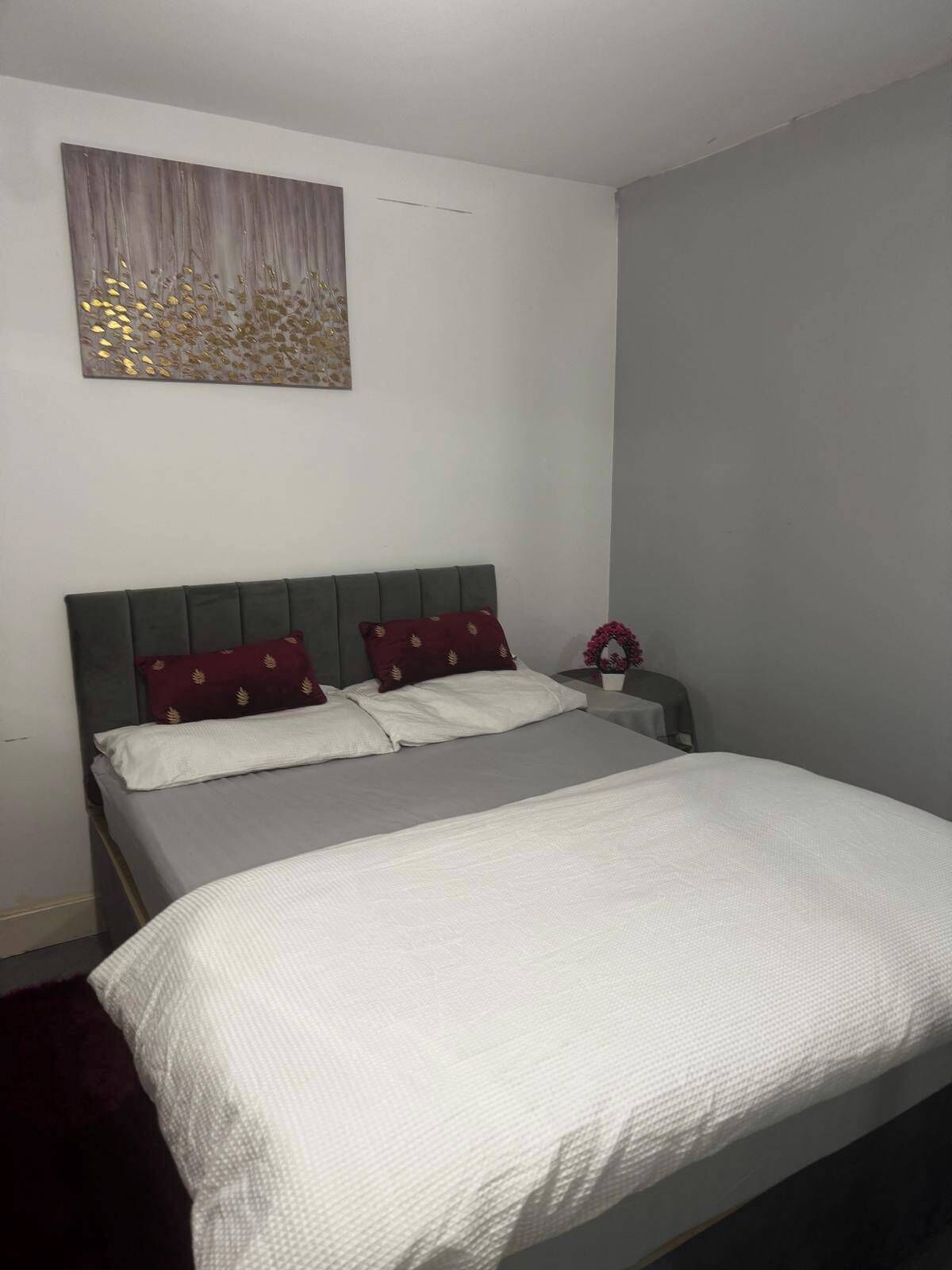 Una cama bien hecha, con cabecera gris y ropa de cama blanca, una mesita de noche con un jarrón de flores y una obra de arte abstracta colgada en la pared.