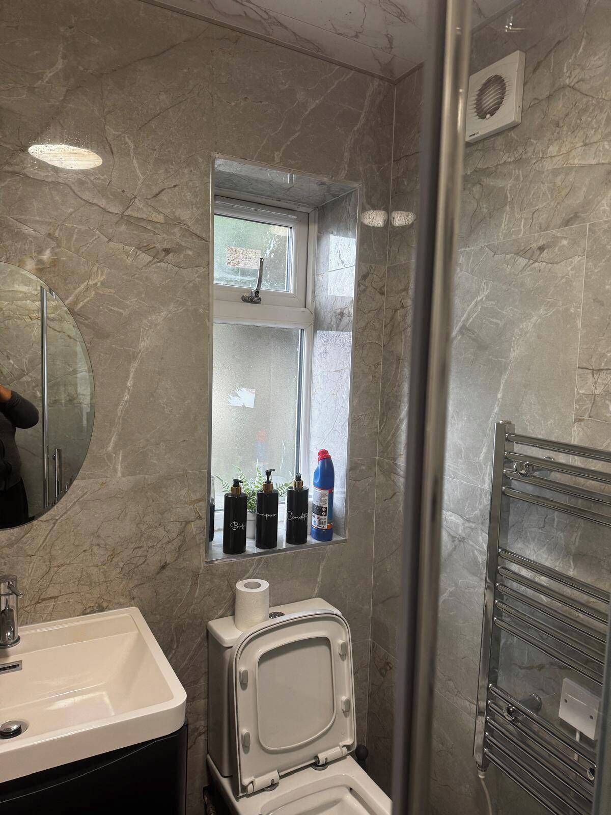 A bathroom with a marble wall, a toilet, a sink, a mirror, and a window with toiletries.