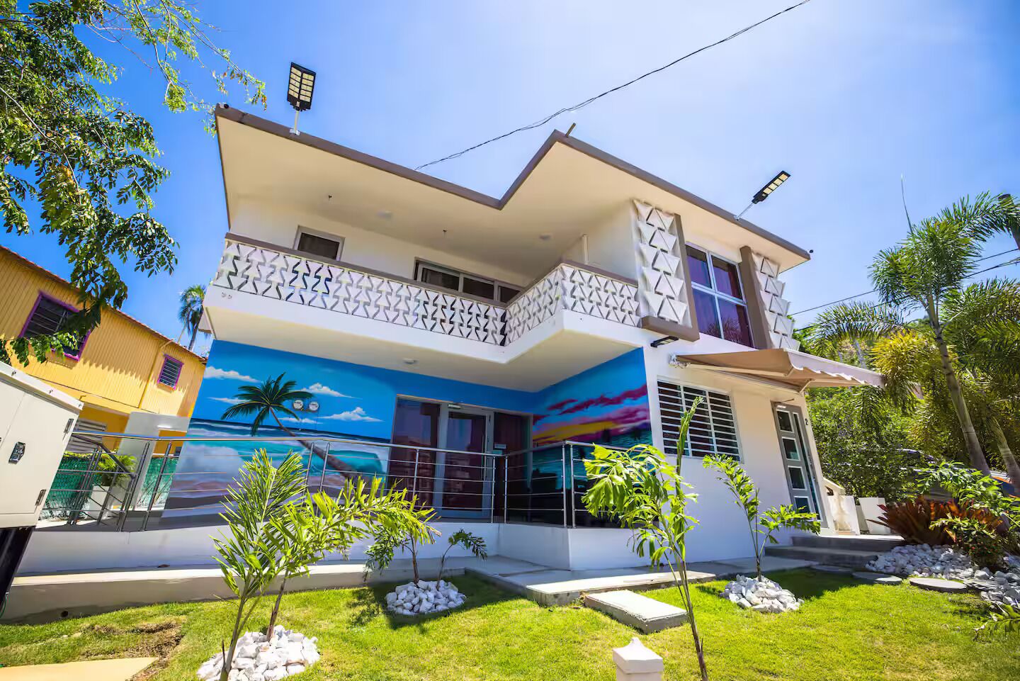 A two-story house with a balcony, a mural of a beach scene, and a well-maintained garden with small trees and rocks.
