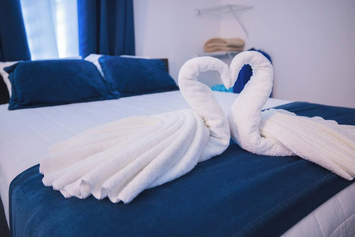 A hotel room with a bed, blue bedding, and two white towels arranged in a heart shape on the bed.