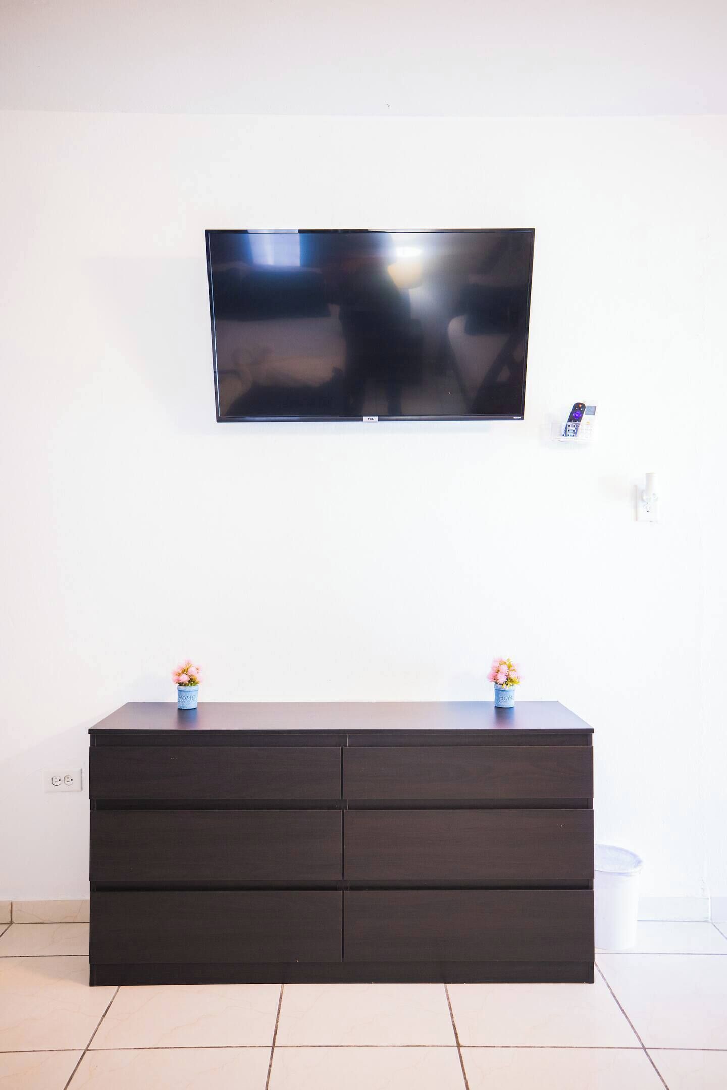 A modern living room with a flat-screen TV mounted on the wall, a dark wooden cabinet with two flower vases, and a small electronic device on a wall-mounted charging station.