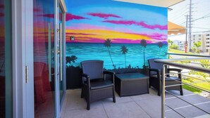 A balcony with a mural of a sunset over the ocean, two wicker chairs, and a table. - Chris Home for 20 Guests - Private Pool & Jacuzzi (Ensenada)