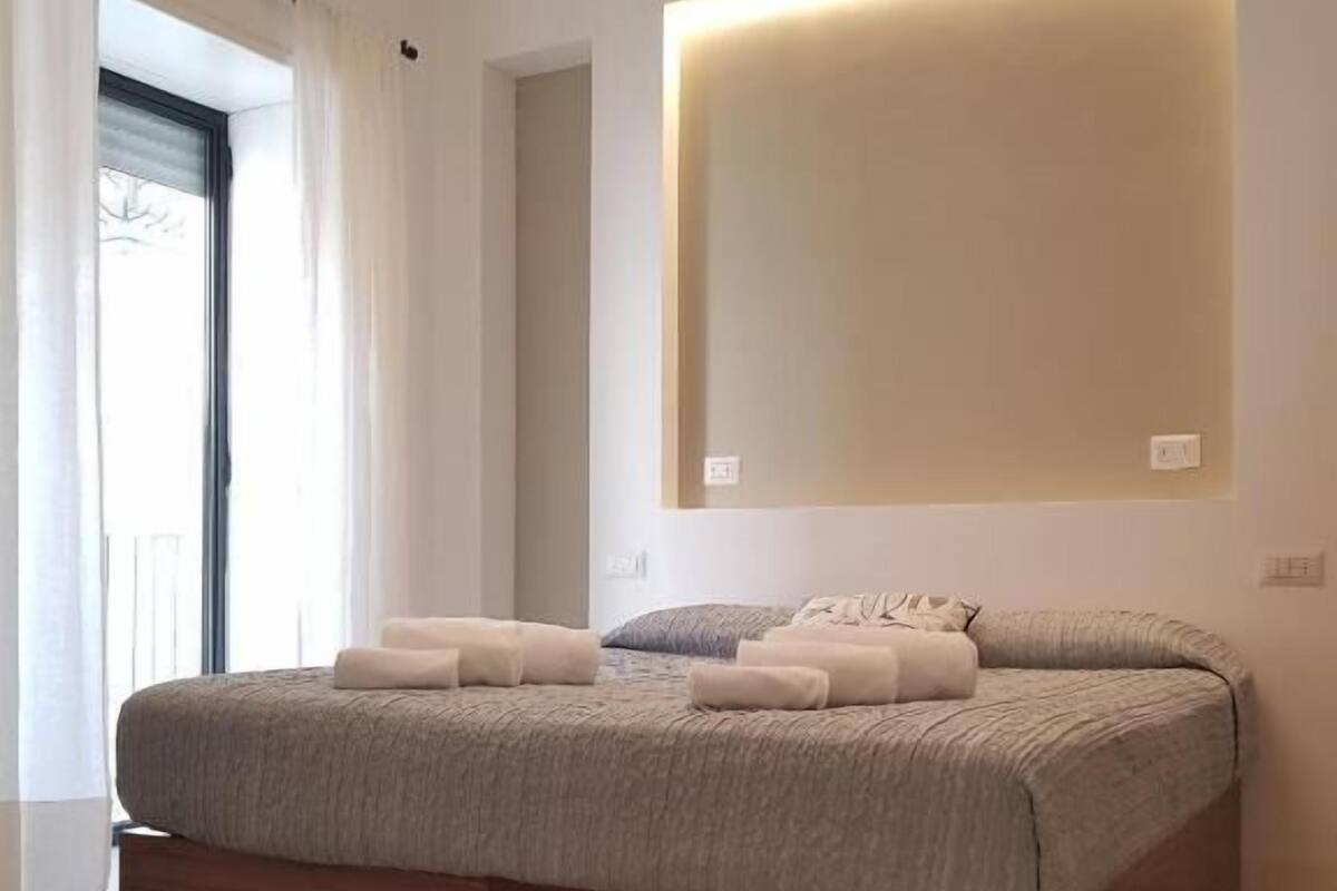 A modern bedroom with a large bed, a wall-mounted light, and a view through a window with curtains.