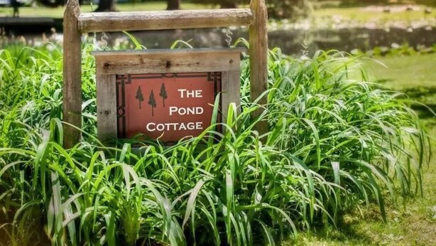 A wooden sign with the text "The Pond Cottage" surrounded by lush green grass. - The Pond Cottage at Rustic River (Austinburg)