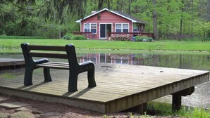A wooden dock with a bench, a red house with a porch, and a pond in the background. - The Pond Cottage at Rustic River (Austinburg)