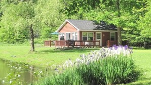 A small house with a deck, surrounded by lush greenery and a pond. - The Pond Cottage at Rustic River (Austinburg)