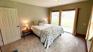 Een slaapkamer met een bed, nachtkastje, lamp en uitzicht op het omliggende landschap door een raam.