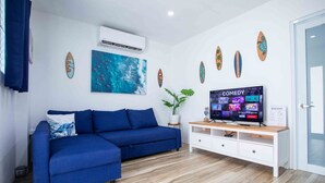 A modern living room with a blue sofa, a flat-screen TV displaying a comedy channel, and decorative surfboards on the wall. - Casa Rosado @ Rincon Rayo Suite Mountain View Pool (Cruces)