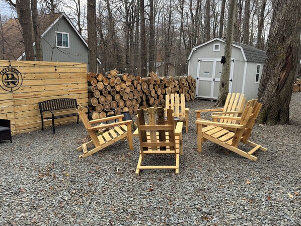 A wooden outdoor seating area with Adirondack chairs, a bench, and a pile of firewood.