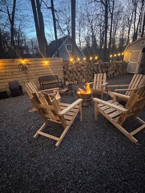A cozy outdoor seating area with Adirondack chairs around a fire pit, a wooden wall with a logo, and a stack of firewood.