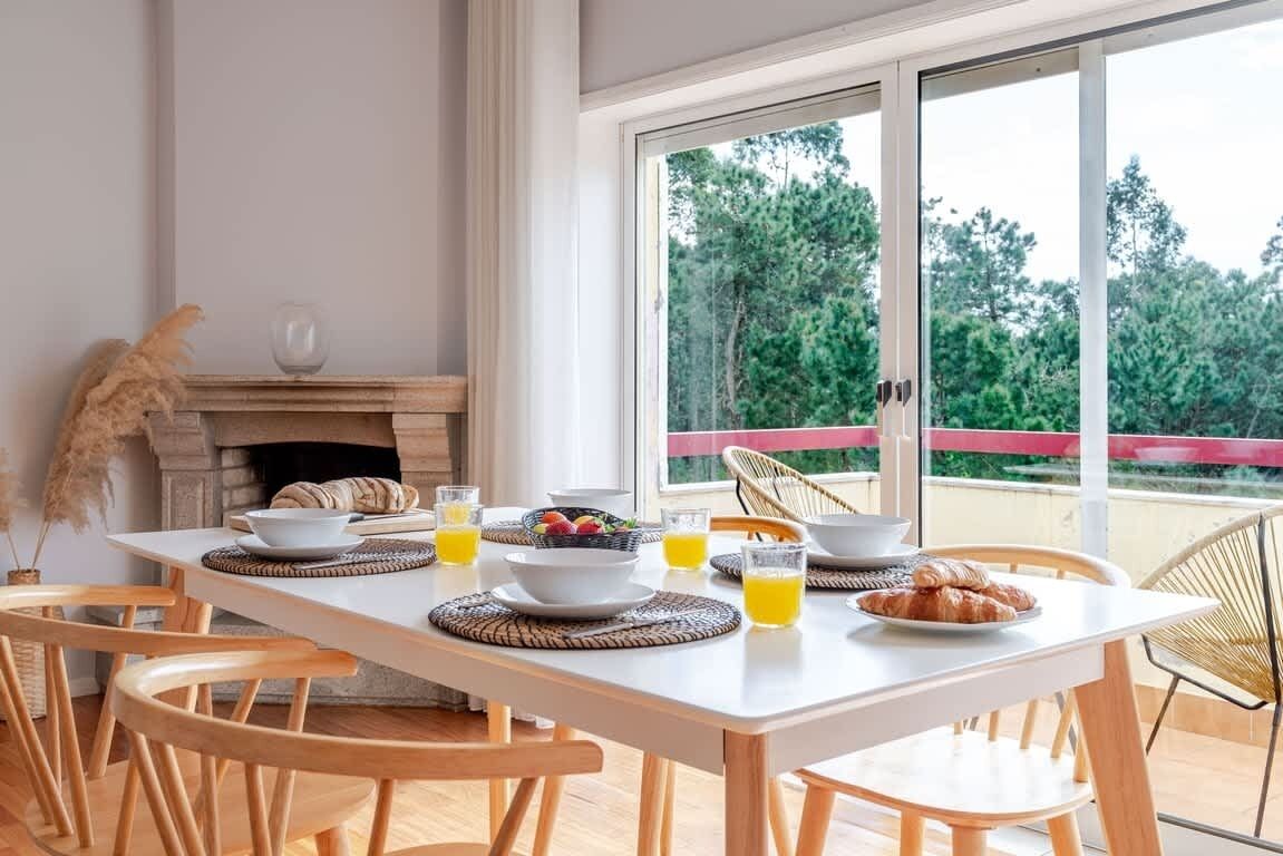 Espaço de refeições com mesa posta para o café da manhã, lareira e vista para árvores por grandes janelas.
