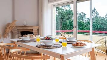 Espaço de refeições com mesa posta para o café da manhã, lareira e vista para árvores por grandes janelas.