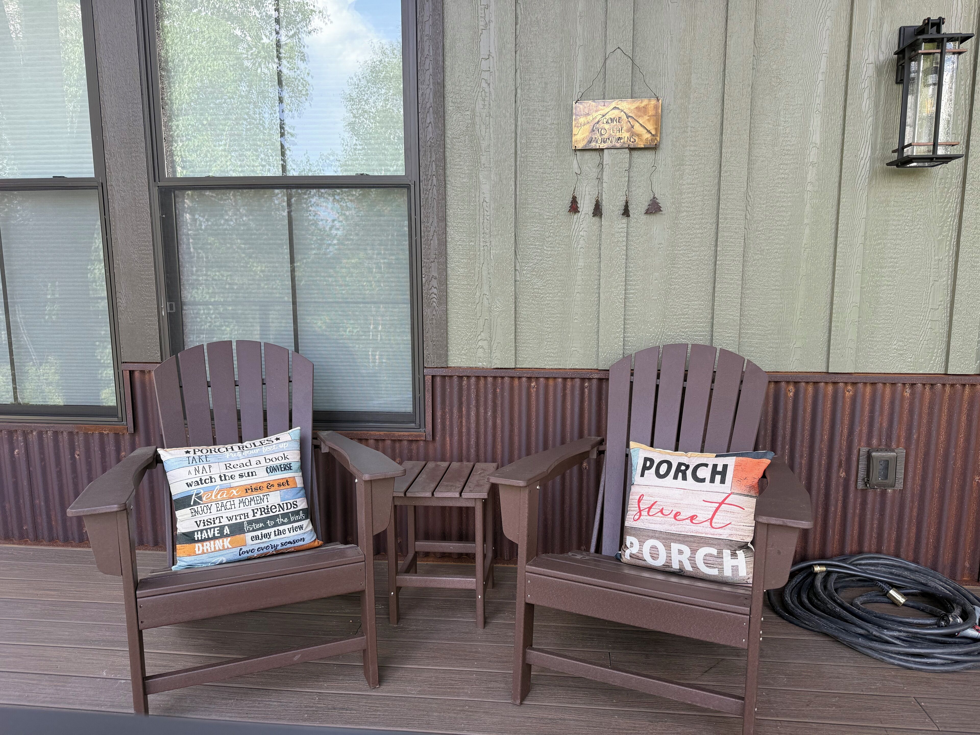 Two Adirondack chairs with pillows, a small table, and a hanging lantern on a wooden porch.