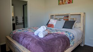 Ein ordentlich bezogenes Bett mit einem blumigen Überwurf, einer violetten Decke und mehreren Kissen. Darüber hängt ein Bild an der Wand.