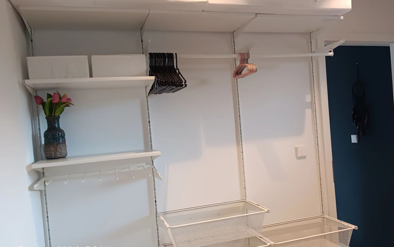 A white storage unit with shelves and hanging baskets, a vase with flowers, and a metal basket.
