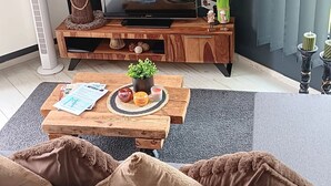 A modern living room with a dark wall, a wooden entertainment unit, a flat-screen TV, a round wall clock, and a cozy seating area with a gray sofa and brown cushions.