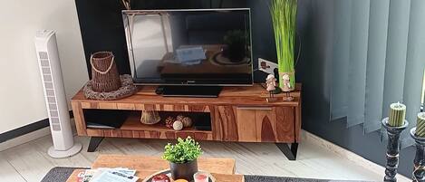 A modern living room with a dark wall, a wooden entertainment unit, a flat-screen TV, a round wall clock, and a cozy seating area with a gray sofa and brown cushions.