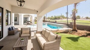 A covered outdoor area with a wicker sofa, a small table, and a view of a swimming pool and landscaped garden.