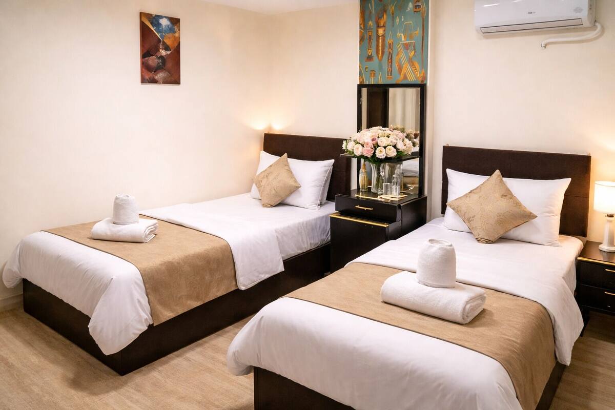 Habitación de hotel con dos camas, cada una con cubrecama blanco, almohadas color beige y una toalla. Se observa un mesita de noche con un florero y un espejo.