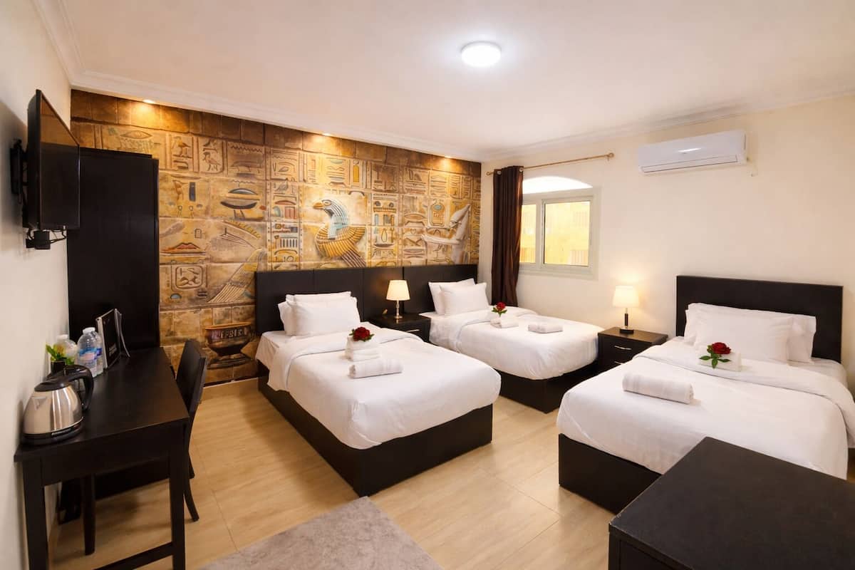 Habitación de hotel con tres camas, un escritorio, un televisor y una pared decorada con diseños del Antiguo Egipto.