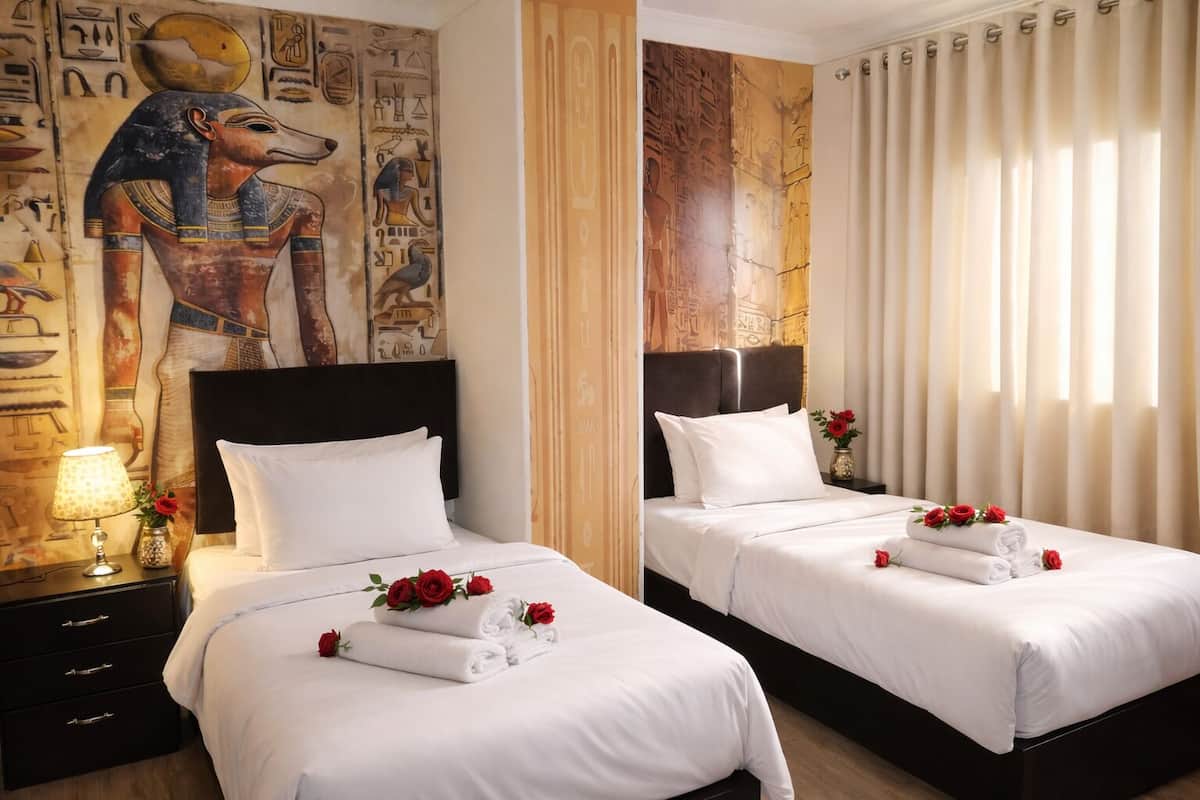 Habitación de hotel con dos camas, una mesita de noche con lámpara y una pared decorada con arte del antiguo Egipto.