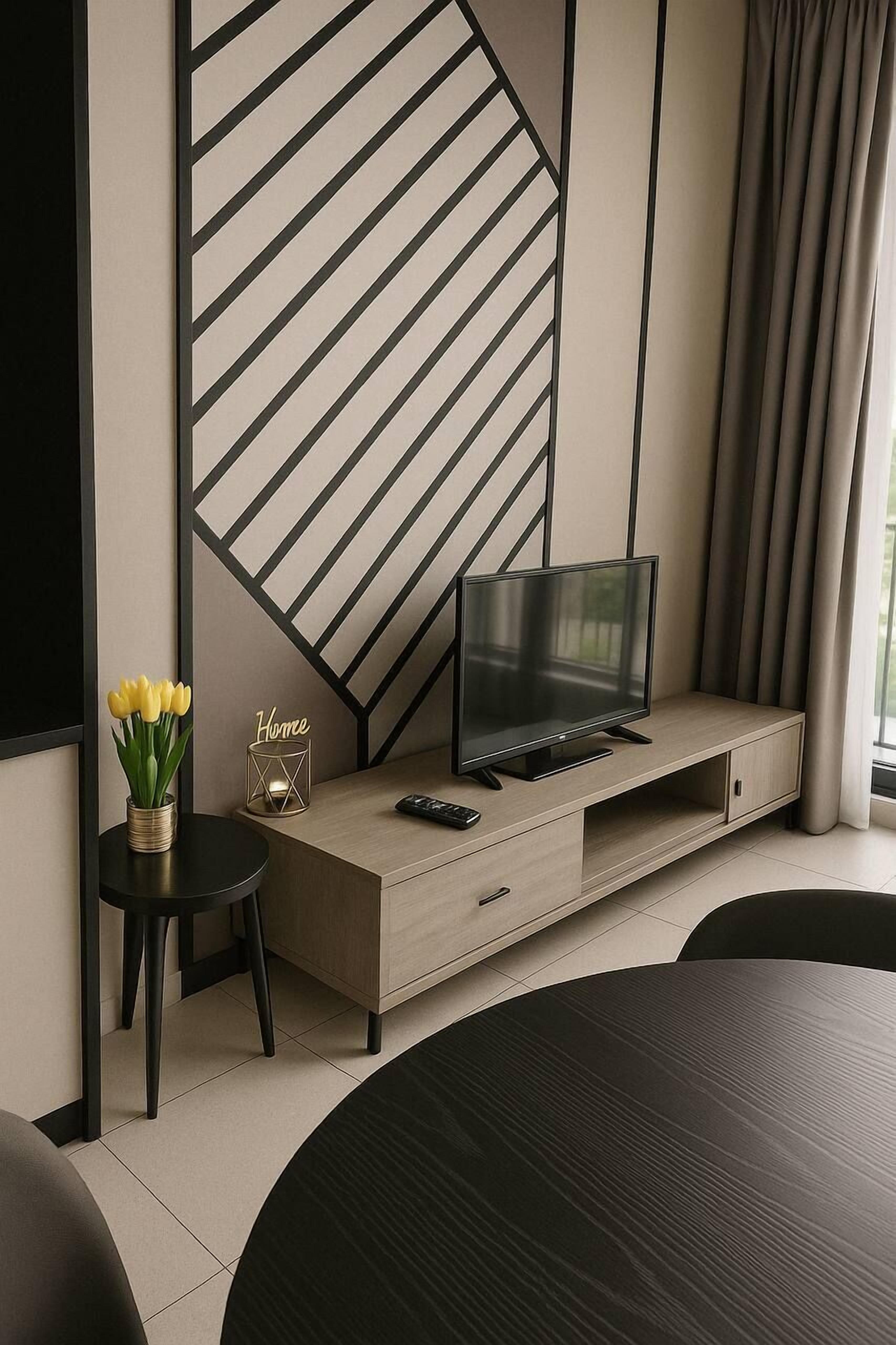 A modern living room with a TV on a wooden console, a small side table with a vase of yellow flowers, and a "Home" sign.