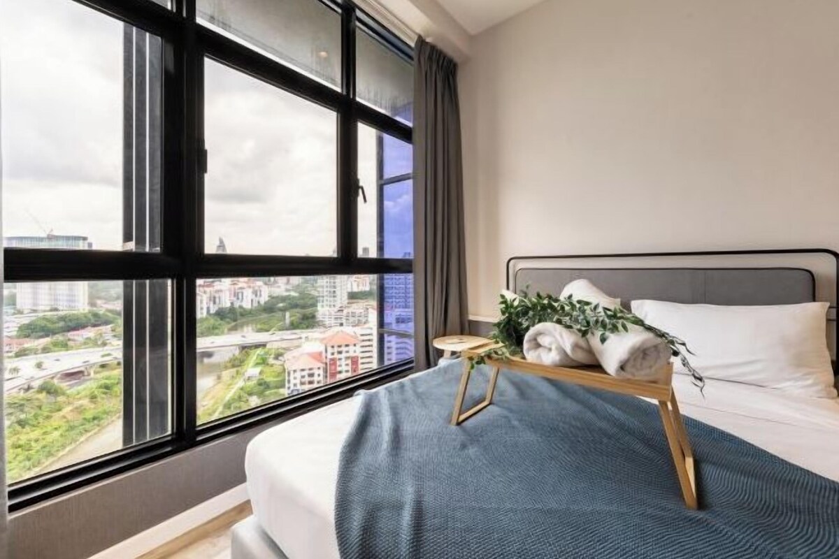 Une chambre d’hôtel avec une grande fenêtre, un lit recouvert d’une couverture bleue, une petite table en bois avec des serviettes et une plante, et une vue sur le paysage urbain.