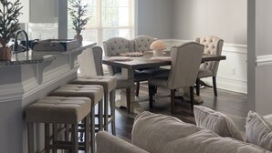 Et moderne spiseområde med et stort bord, flere stole, en bardisk med skamler og en lysekrone over det.