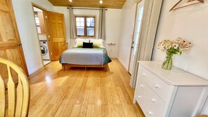 A bedroom with a wooden floor, a bed with a striped bedspread, a dresser, a chair, and a window with curtains.