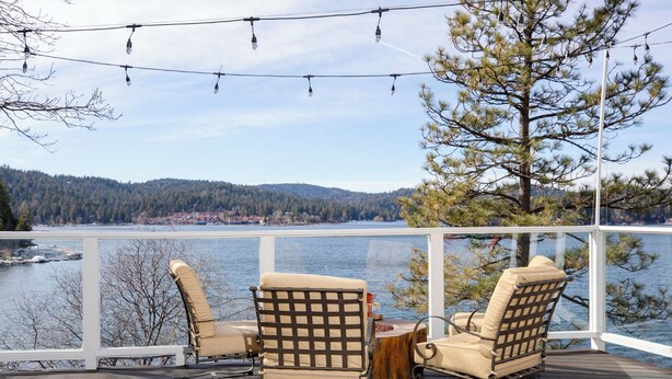 A deck with two rocking chairs facing a lake, a wooden table, and string lights. - Stunning Lake Views To Die For On Lake Arrowhead (Lake Arrowhead)