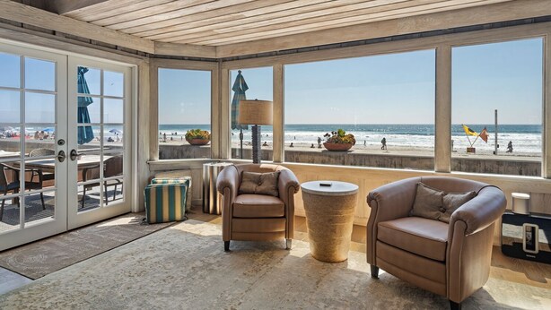 A sunlit room with large windows overlooking a beach, featuring leather sofas, a round table, and a potted plant.