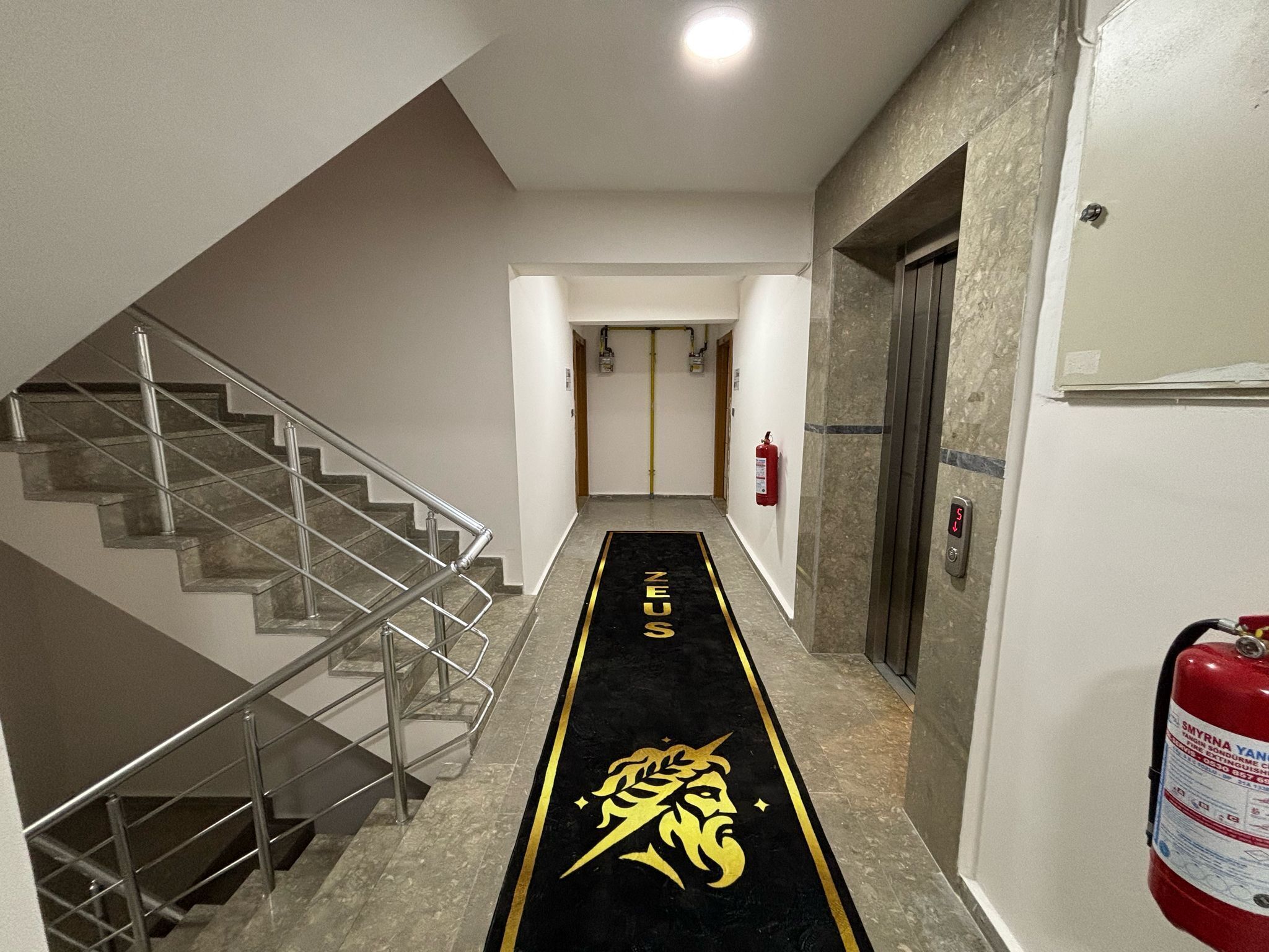 A hotel hallway with a black carpet featuring a golden lion's head emblem, a staircase on the left, and a fire extinguisher on the right.