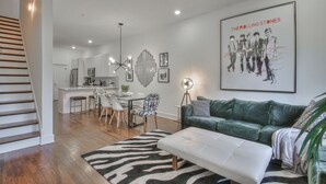 Ein modernes Wohnzimmer mit einem grünen Sofa, einem weißen Hocker und einem Teppich mit Zebra-Muster. Links befindet sich eine Treppe, ein Essbereich mit Tisch und Stühlen sowie eine Küche im Hintergrund.