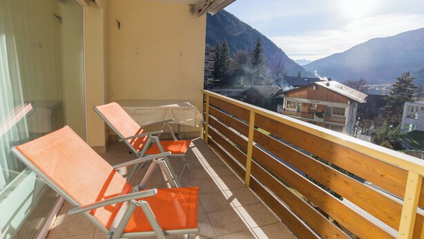 A balcony with orange chairs, a table, and a view of mountains and buildings. - The Cristal apartment house is situated in a quiet, sunny and central location (Leukerbad)