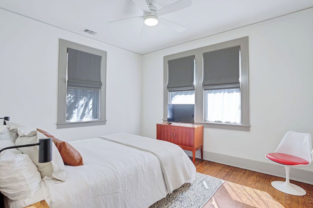 A bedroom with a bed, a wooden dresser, a red chair, and two windows with blinds.