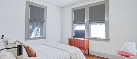 A bedroom with a bed, a wooden dresser, a red chair, and two windows with blinds.