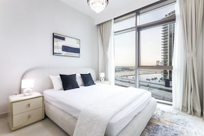 A hotel room with a large bed, a nightstand, a lamp, a framed picture on the wall, and a view of a cityscape through a large window.