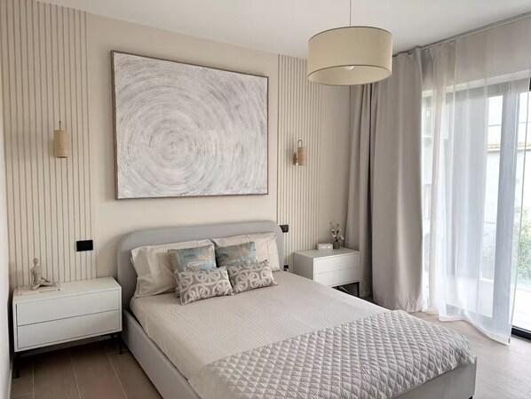 Un dormitorio moderno con una cama grande, mesitas de noche, una lámpara de pared y un ventanal con cortinas.