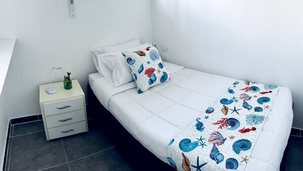 A neatly made bed with a white duvet and a decorative pillow, a bedside table with a lamp and a vase, and a window with a view of greenery. - Turbay House 13 (Cartagena de Indias)