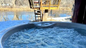 Un jacuzzi con banca de madera y escalera, rodeado por un paisaje natural con árboles y una masa de agua.